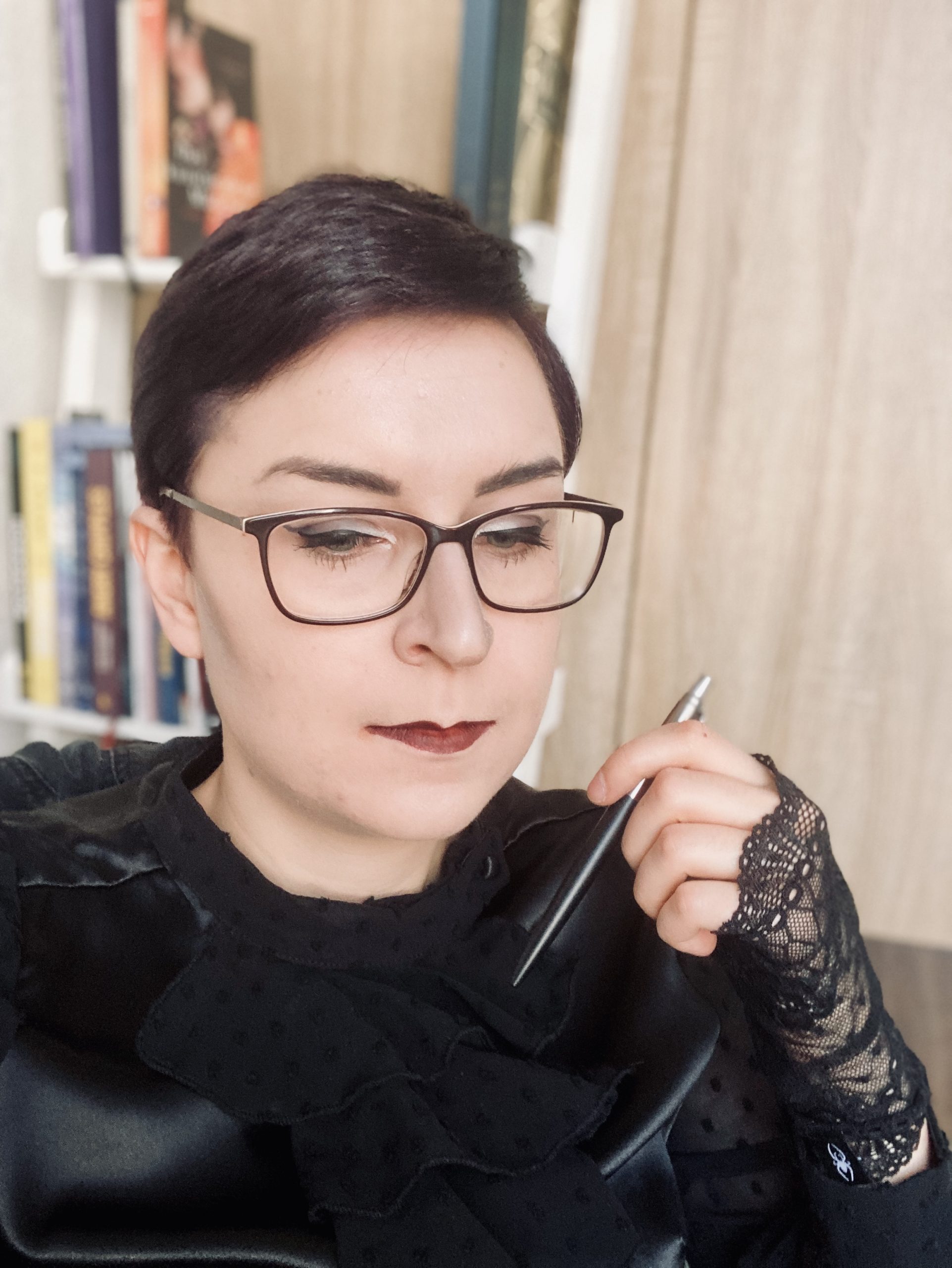 Bild von Charline Winter: Eine weiße Person mit Brille und kurzen Haaren in einer schwarzen Bluse, die einen Stift in der Hand hält und nachdenklich nach unten schaut. Im Hintergrund ein Bücherregal.